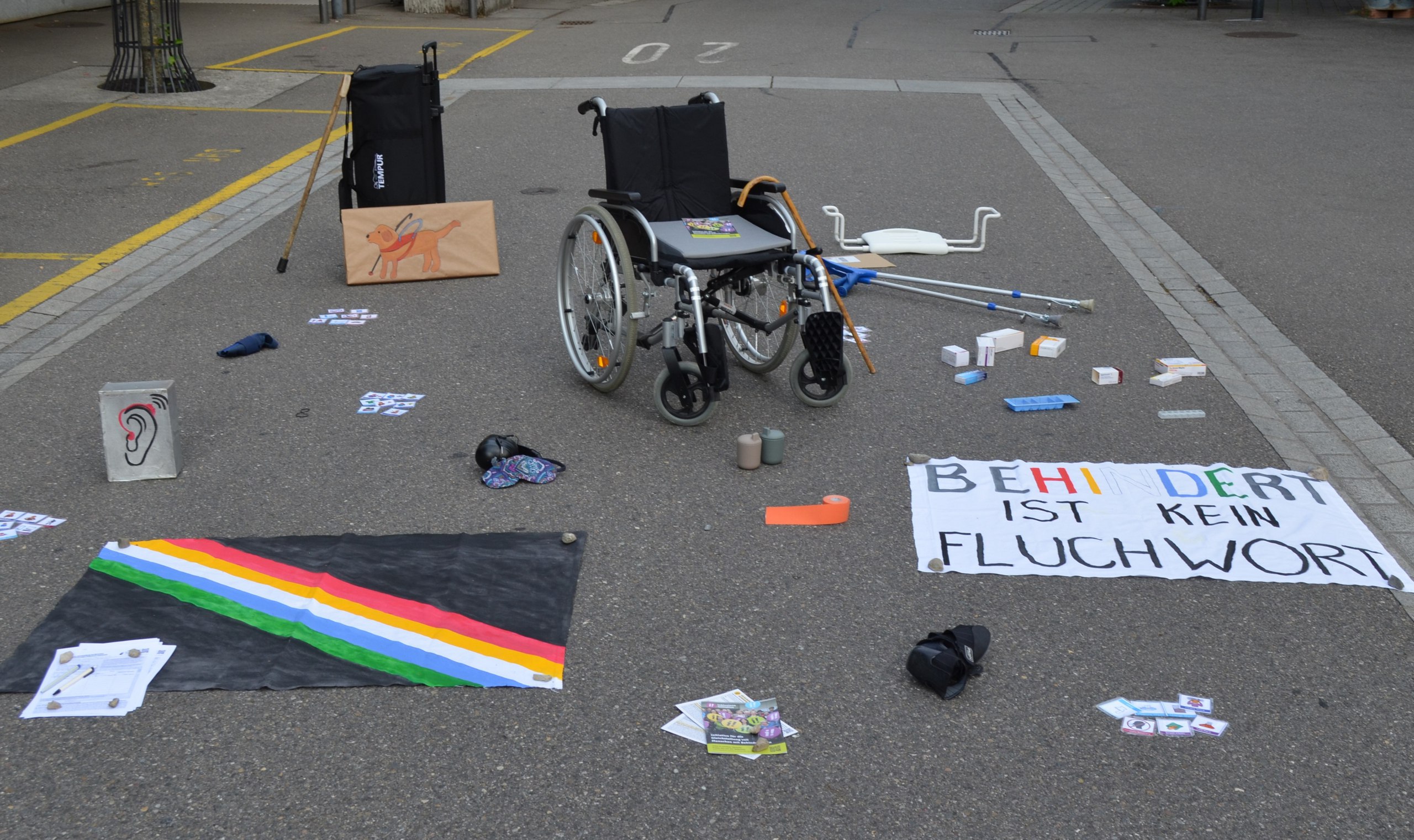 "Behindert ist kein Fluchwort" steht auf einem Transparent neben verschiedensten Hilfsgegenständen, die auf der Strasse verteilt liegen.